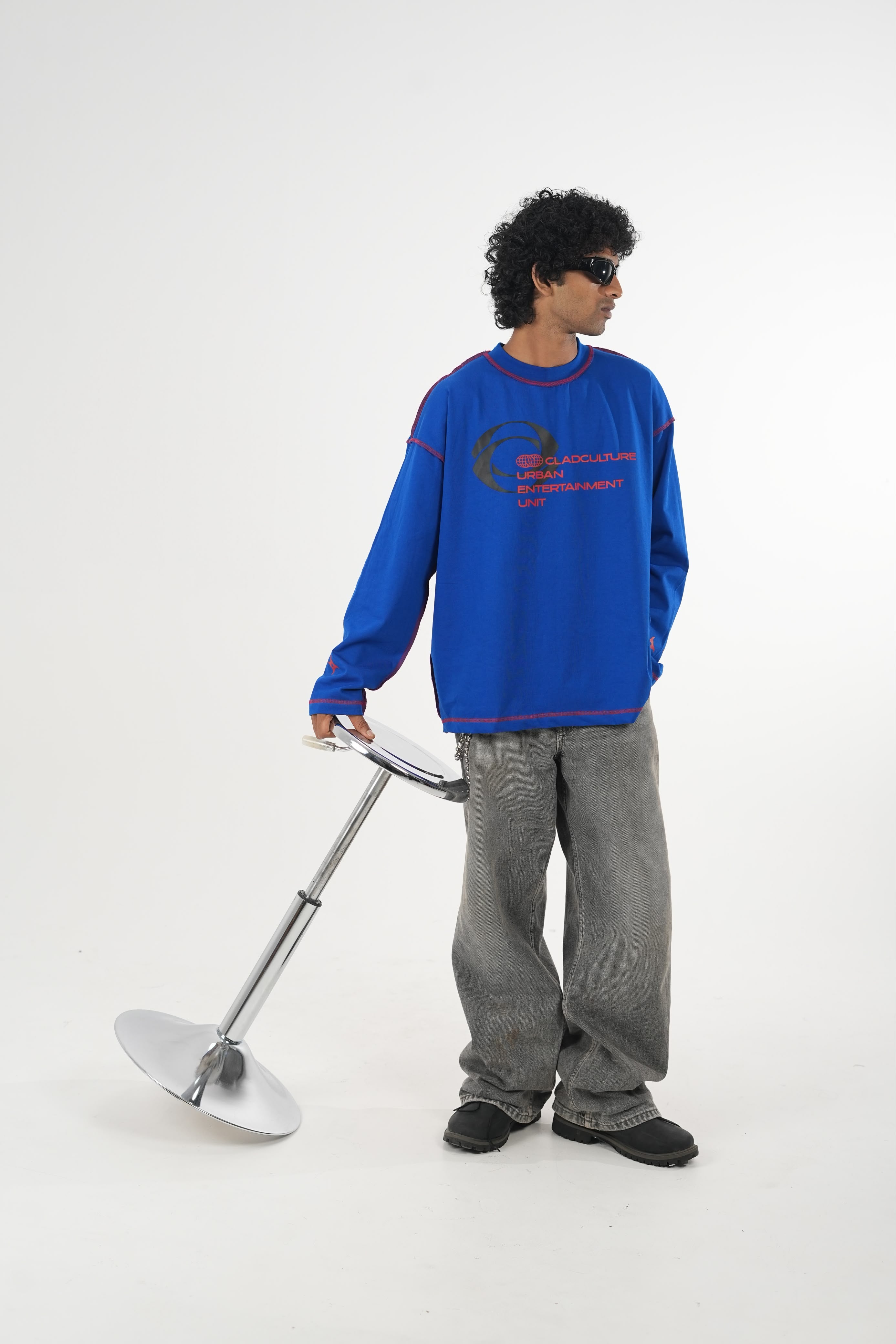 Model wearing a royal blue oversized long sleeve graphic T-shirt with contrast stitching and Urban Entertainment front print, paired with baggy grey jeans in a minimalist studio setting.
