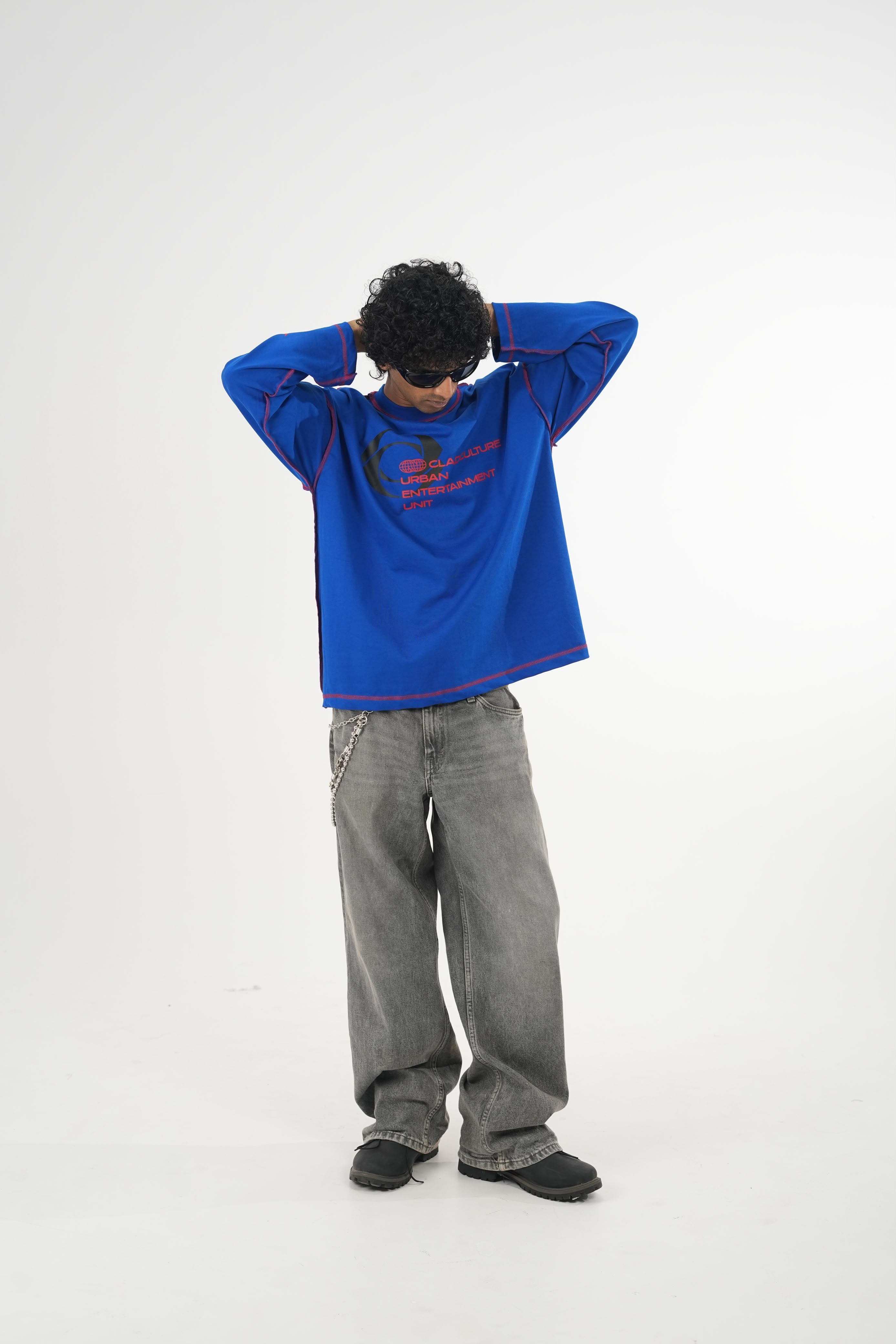 Model wearing a royal blue oversized long sleeve graphic T-shirt with contrast stitching and Urban Entertainment front print, paired with baggy grey jeans in a minimalist studio setting.