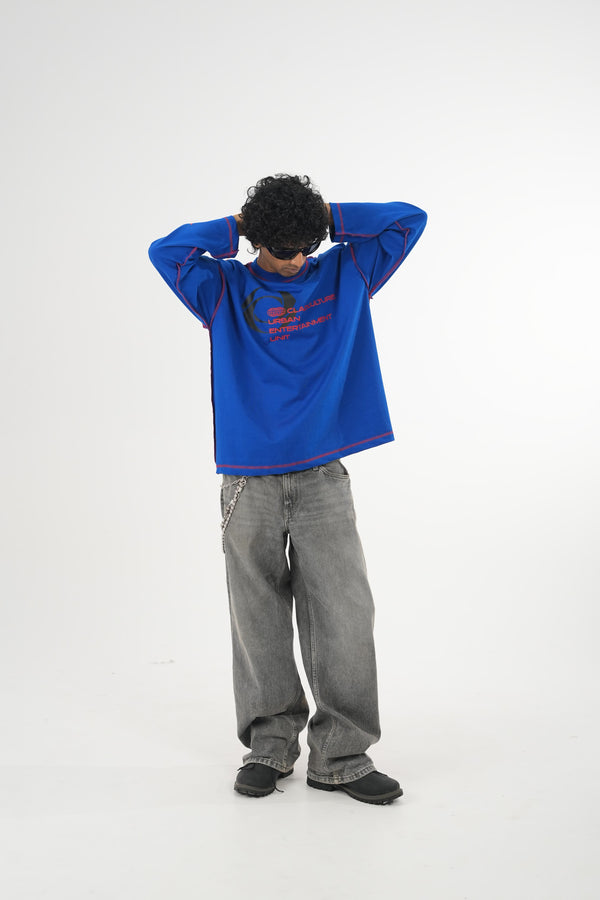 Model wearing a royal blue oversized long sleeve graphic T-shirt with contrast stitching and Urban Entertainment front print, paired with baggy grey jeans in a minimalist studio setting.