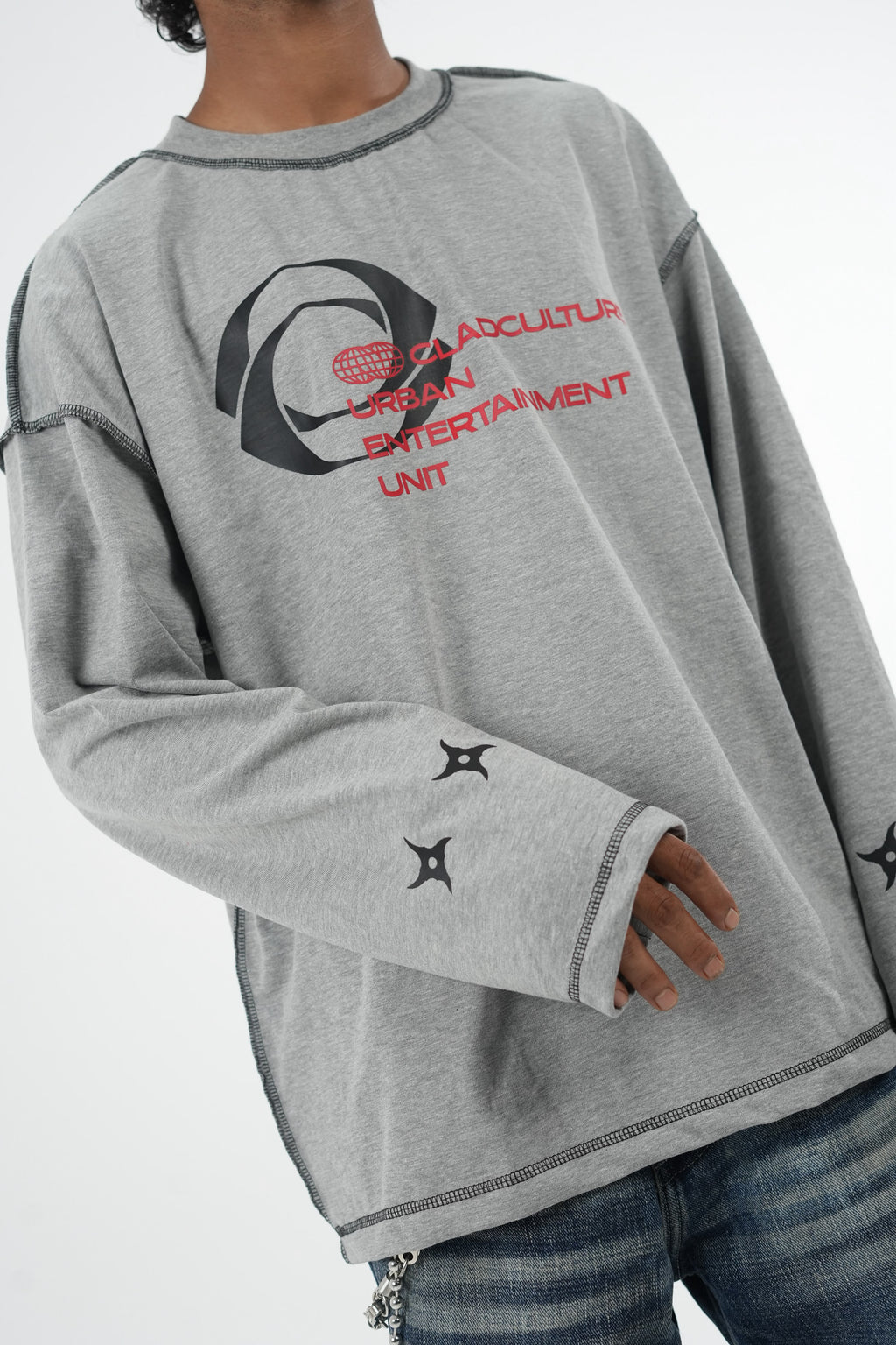 Model wearing a grey oversized long sleeve graphic T-shirt with contrast stitching and Urban Entertainment front print, paired with baggy grey jeans in a minimalist studio setting.
